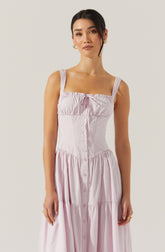 A woman is wearing a sleeveless, light pink sundress with wide straps that have lace details. The dress features a fitted bodice with gathered fabric at the top, a row of buttons running down the front, and a gathered, tiered skirt.